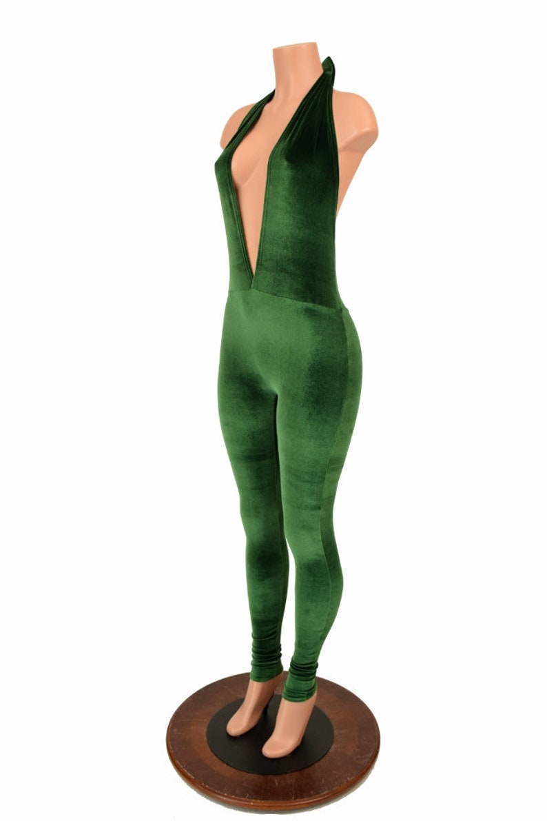 May include: A green velvet jumpsuit with a plunging neckline and halter top design. The one-piece garment features a form-fitting silhouette and long legs. The jumpsuit is displayed on a mannequin with a wooden base.