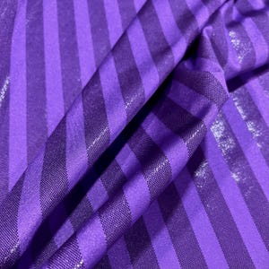 May include: A vibrant purple fabric featuring a repeating pattern of alternating vertical stripes. The design consists of one set of smooth, solid purple stripes paired with another set of textured, shimmering purple stripes that catch the light.