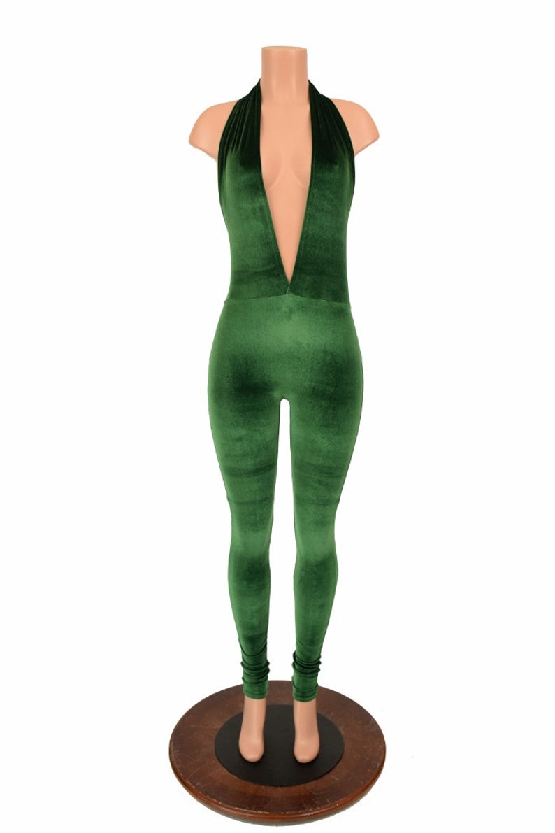 May include: A green velvet jumpsuit with a halter neckline and a plunging V-neck. The jumpsuit is displayed on a mannequin and has a form-fitting silhouette. The legs are long and fitted, with a slight gathering at the ankles.