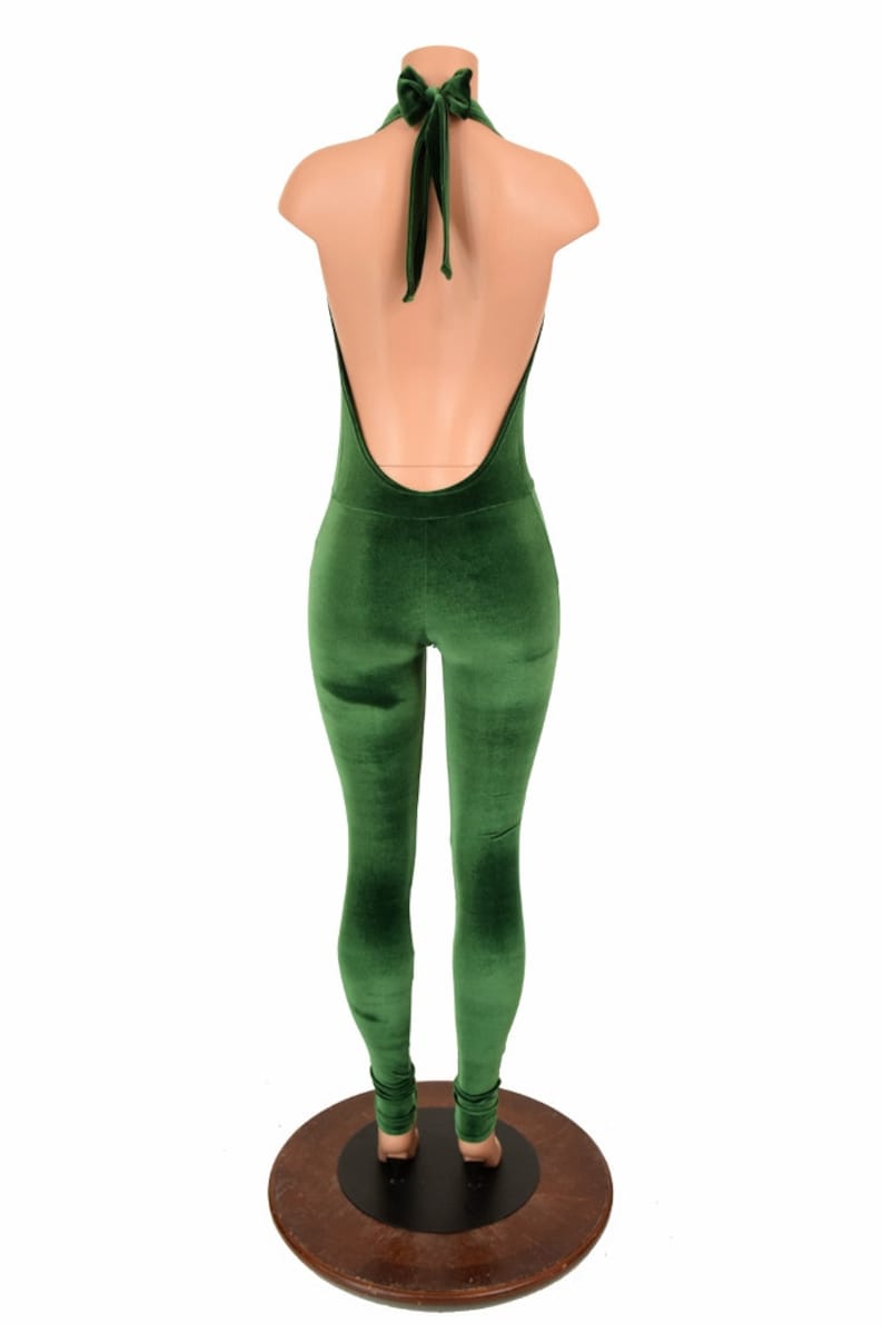 May include: A green velvet jumpsuit with a halter neck and open back. The jumpsuit is displayed on a mannequin, highlighting its close-fitting design. The fabric has a soft, plush texture, and the colour is a deep, rich green.