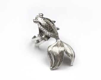 May include: A silver ring featuring a detailed, three-dimensional design of a goldfish. The fish is facing upwards with its tail curled around the ring band.