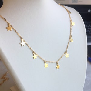 May include: A gold-colored necklace with star-shaped pendants. The necklace is displayed on a white mannequin. The stars are evenly spaced along the chain, creating a delicate and celestial design. The necklace is a simple yet elegant piece of jewelry.
