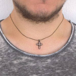 May include: A dark metal cross pendant necklace. The cross is a simple, outlined design, suspended from a delicate chain. The necklace is worn against a gray t-shirt.