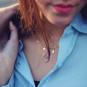 May include: A gold-colored necklace with star-shaped pendants is worn over a light blue button-down shirt. A tan coat is draped over the shoulders. The image is a close-up, focusing on the necklace and shirt.