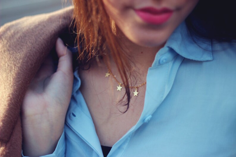 May include: A gold-colored necklace with star-shaped pendants is worn over a light blue button-down shirt. A tan coat is draped over the shoulders. The image is a close-up, focusing on the necklace and shirt.