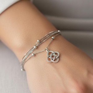 May include: Silver bracelet featuring three delicate strands and small beaded accents. A Celtic knot charm dangles from the bracelet. The jewelry is designed to be worn on the wrist.