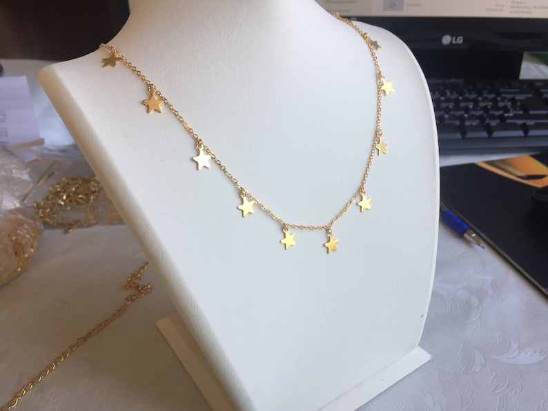 May include: A gold-colored necklace with star-shaped pendants. The necklace is displayed on a white mannequin. The stars are evenly spaced along the chain, creating a delicate and celestial design. The necklace is a simple yet elegant piece of jewelry.