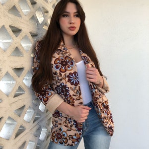 May include: A woman wearing a beige blazer with a floral pattern in shades of brown, blue, and white. She is also wearing blue jeans and a white tank top.