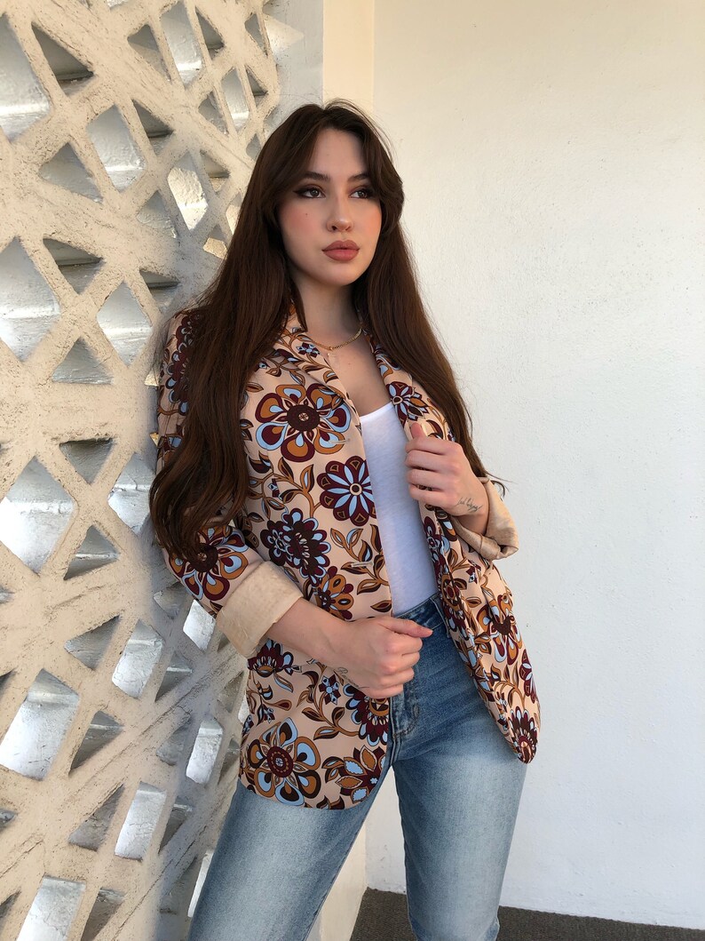 May include: A woman wearing a beige blazer with a floral pattern in shades of brown, blue, and white. She is also wearing blue jeans and a white tank top.