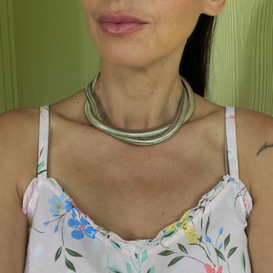 May include: A silver double-layered snake chain necklace. The necklace is worn by a person with a pink floral top.