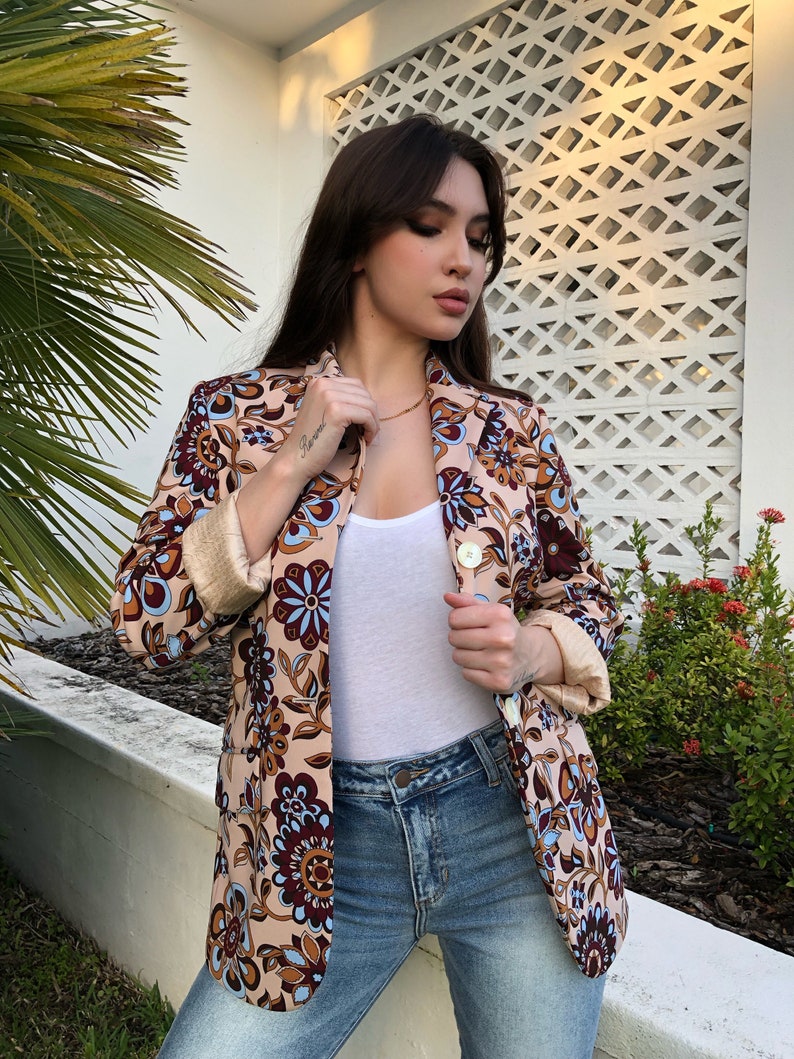 May include: A woman wearing a brown blazer with a floral pattern. The blazer has a light blue and brown floral design. The woman is also wearing a white tank top and blue jeans.
