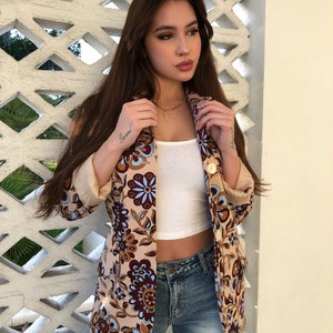 May include: A woman wearing a beige blazer with a floral pattern in shades of brown, blue, and white. The blazer has a gold button closure and a white tank top underneath. She is also wearing blue jeans.