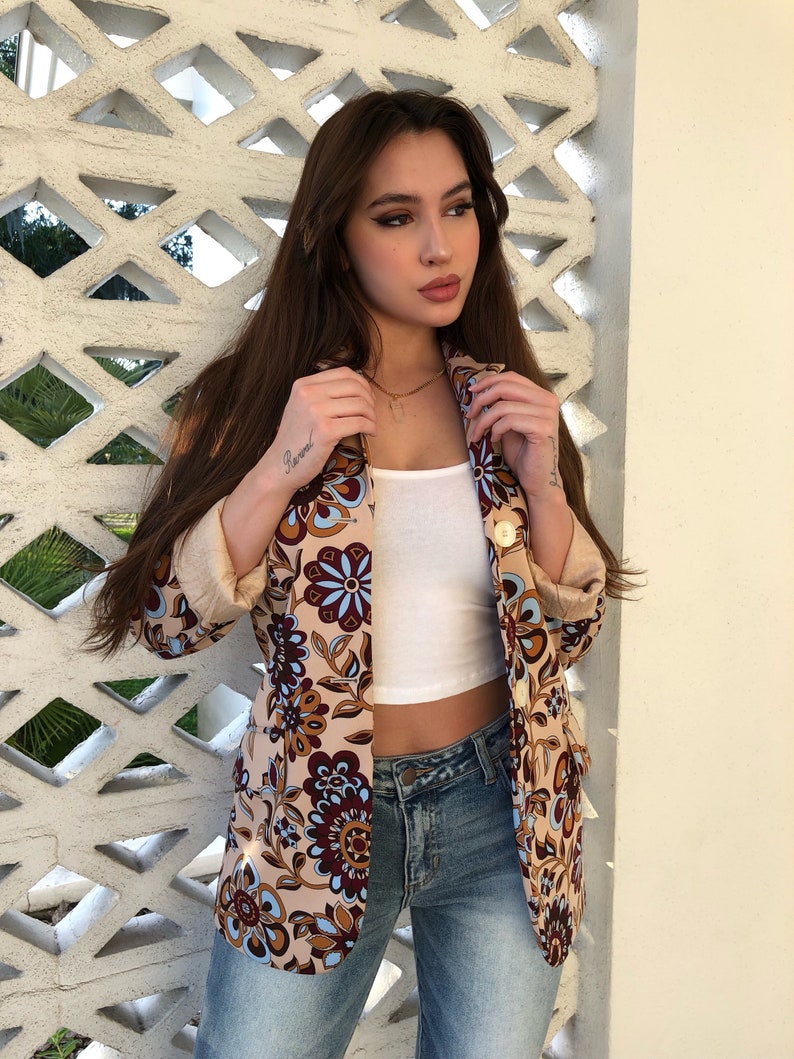 May include: A woman wearing a beige blazer with a floral pattern in shades of brown, blue, and white. The blazer has a gold button closure and a white tank top underneath. She is also wearing blue jeans.
