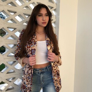 May include: A woman wearing a beige blazer with a floral pattern in shades of brown, blue, and white. She is also wearing a white tank top and blue jeans.