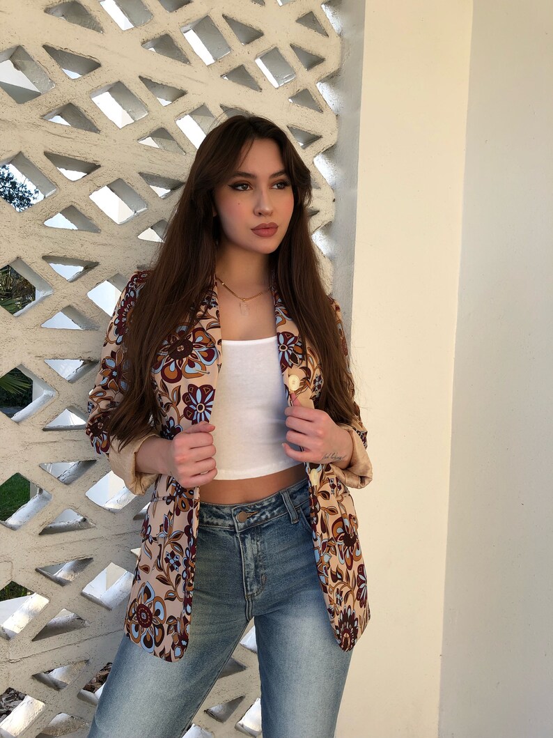 May include: A woman wearing a beige blazer with a floral pattern in shades of brown, blue, and white. She is also wearing a white tank top and blue jeans.