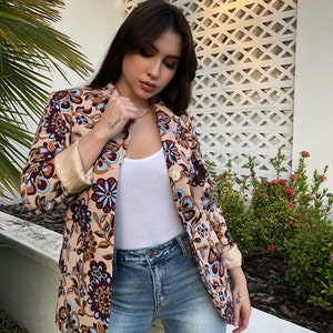 May include: A woman wearing a beige blazer with a floral pattern in shades of brown, blue, and white. The blazer has a gold button and a white collar. She is also wearing a white tank top and blue jeans.