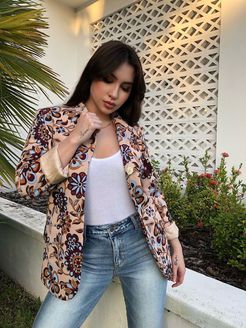 May include: A woman wearing a beige blazer with a floral pattern in shades of brown, blue, and white. The blazer has a gold button and a white collar. She is also wearing a white tank top and blue jeans.
