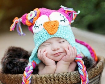 May include: A baby wearing a crocheted owl hat with pink, blue, orange, and white yarn. The hat has long braids that are also crocheted. The baby is lying in a brown wicker basket.