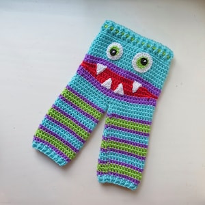 May include: Crocheted baby pants with a monster face design. The pants are blue, green, purple, and red with white teeth and eyes.