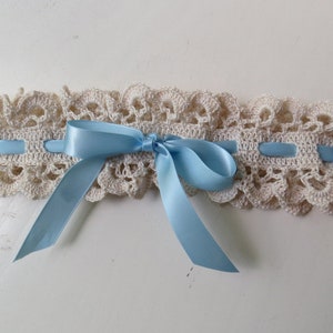 May include: Ivory crochet garter with a scalloped edge and light blue satin ribbon. The garter features a delicate, openwork design and a bow in the center. A classic accessory for a special occasion.