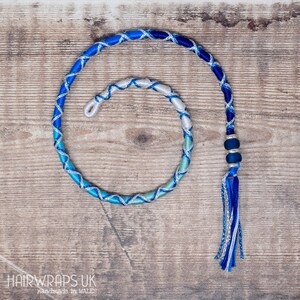 May include: A blue and silver hair wrap with a tassel and beads. The wrap is made of braided cords and is coiled into a circle. The background is a wooden surface.