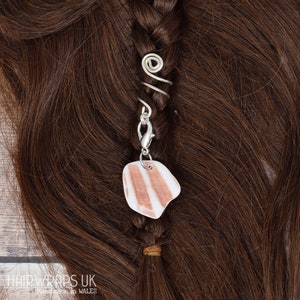 Puede incluir: Un envoltorio de pelo plateado con un diseño en espiral y un amuleto de piedra rosa y blanca. El envoltorio de pelo está sujeto a una trenza de pelo castaño.