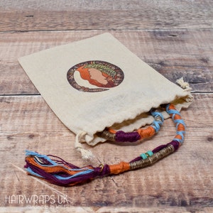 May include: A colorful hair wrap with a tassel, made with multiple strands of yarn in shades of orange, blue, purple, and brown. The wrap is tied in a circle and has small turquoise beads. The wrap is displayed in a small, white drawstring bag with a logo that says "Hairwraps UK Handmade in Wales".