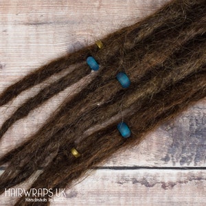 May include: Brown dreadlocks with blue and gold beads. The beads are cylindrical and have a smooth, shiny finish. The dreadlocks are arranged in a fan shape on a wooden surface.