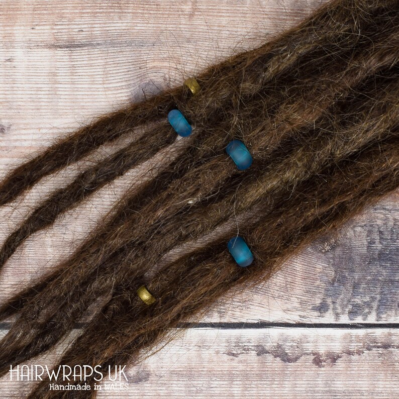 May include: Brown dreadlocks with blue and gold beads. The beads are cylindrical and have a smooth, shiny finish. The dreadlocks are arranged in a fan shape on a wooden surface.