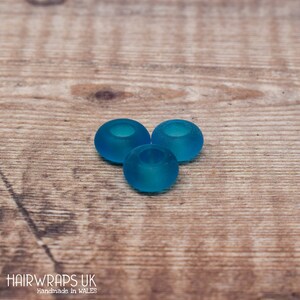 May include: Three teal blue glass beads with a hole in the centre. The beads are on a wooden surface.