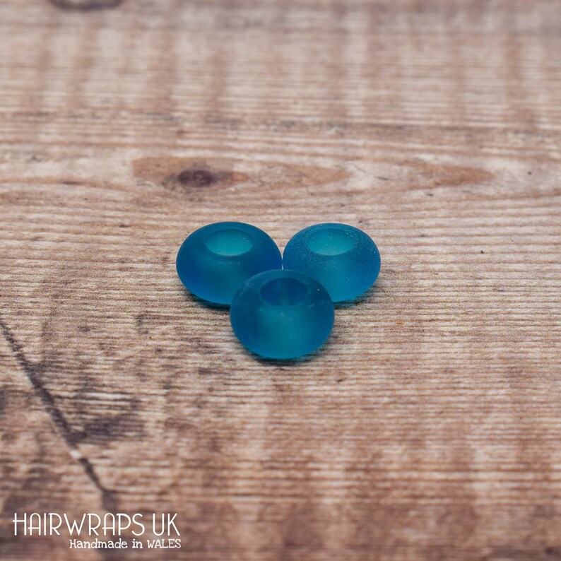 May include: Three teal blue glass beads with a hole in the centre. The beads are on a wooden surface.
