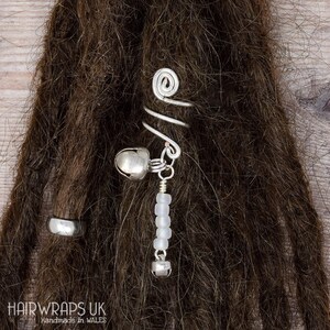 Op de afbeelding: Zilveren dreadlock kralen met een zilveren spiraalvormige haarband en twee zilveren belletjes. De kralen zijn wit en ondoorzichtig. De haarband is spiraalvormig en heeft een kleine lus aan het einde. De belletjes zijn met een klein zilveren kettinkje aan de haarband bevestigd.