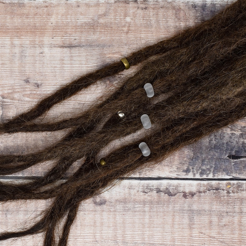 May include: Brown dreadlocks with clear and gold beads. The dreadlocks are on a wooden surface.