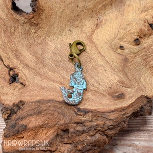 May include: A blue and bronze mermaid charm with a lobster clasp. The charm is on a wooden surface.