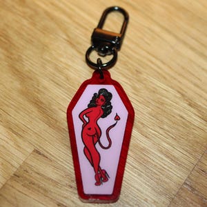 May include: Red and pink keychain featuring a devil woman illustration. The keychain has a black clasp and a coffin-shaped pendant with a red outline. The devil woman is red with black hair and a heart-shaped tail.