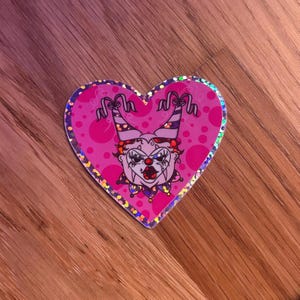 May include: A heart-shaped sticker with a holographic border featuring a cartoon clown face. The clown has a white face, red lips, and a purple and red jester hat. The background is pink with small pink dots.