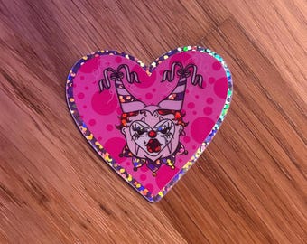 May include: A heart-shaped sticker with a holographic border featuring a cartoon clown face. The clown has a white face, red lips, and a purple and red jester hat. The background is pink with small pink dots.
