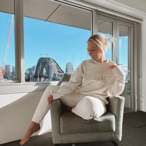 May include: A woman wearing a white sweatshirt with the word "BRIDE" embroidered on the front. She is sitting in a chair in front of a window with a view of a city skyline.