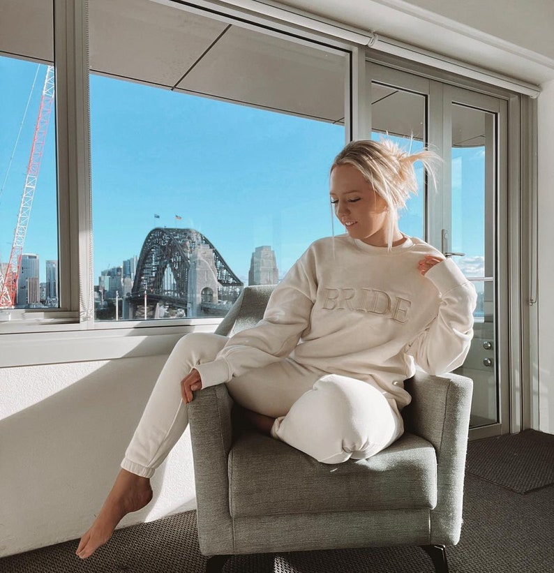 May include: A woman wearing a white sweatshirt with the word "BRIDE" embroidered on the front. She is sitting in a chair in front of a window with a view of a city skyline.