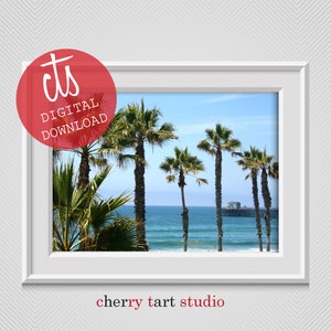May include: A framed print depicting a coastal scene with palm trees, the ocean, and a pier under a blue sky. The image is labelled "DIGITAL DOWNLOAD" in a red circle. The text "cherry tart studio" is at the bottom.