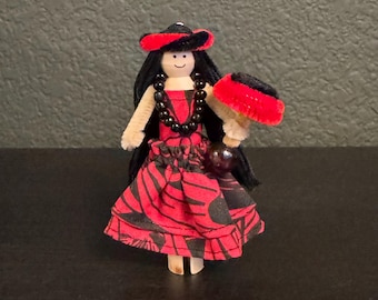 Puede incluir: Muñeca hecha a mano vestida con un vestido rojo y negro estampado, cabello negro y un sombrero rojo. La muñeca tiene un collar de cuentas negras y sostiene un abanico rojo y negro. La muñeca está hecha de madera y limpiapipas.