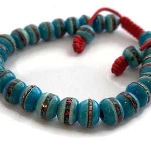 May include: A turquoise blue mala bracelet with red and white accents. The beads are round and have a smooth, polished finish. The bracelet is tied with a red cord.