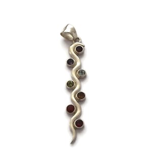 May include: A silver pendant with a flowing, wavy design, adorned with a sequence of colourful gemstones. The gemstones include red, purple, green, and blue stones. The pendant has a small loop at the top for suspension.