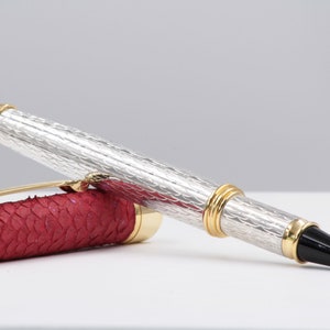 Può includere: Una penna d'argento e oro con un corpo in pelle rossa. La penna ha un fusto argentato testurizzato e una fascia dorata vicino alla punta. Il corpo in pelle ha un motivo squamoso.