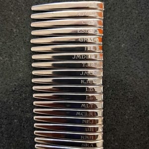 May include: A stack of silver-toned metal collar stays, each featuring a unique engraved monogram including AJC, CJE, ESD, GRAC, HIL, JMDLF, TJE, JMB, KJC, LB, LL, MLT, MCLR, NEJ, JH, RAH, and SRA, arranged in a vertical column on a dark textured surface.