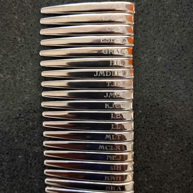 May include: A stack of silver-toned metal collar stays, each featuring a unique engraved monogram including AJC, CJE, ESD, GRAC, HIL, JMDLF, TJE, JMB, KJC, LB, LL, MLT, MCLR, NEJ, JH, RAH, and SRA, arranged in a vertical column on a dark textured surface.