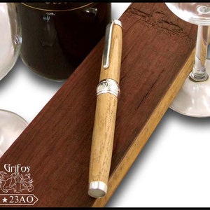 May include: A wooden pen with silver accents and a decorative design. The pen is resting on a wooden surface with two wine glasses.