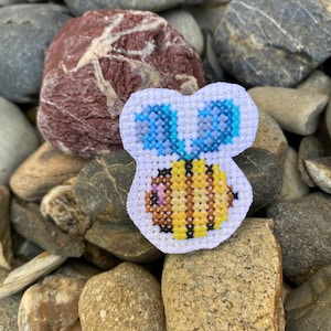 May include: A small, white, embroidered patch featuring a yellow and brown bee with blue wings. The bee has a pink heart on its body.
