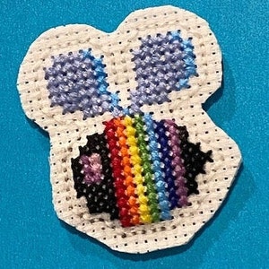 Puede incluir: Un parche de punto de cruz blanco con un diseño de abeja arcoíris. La abeja tiene un cuerpo negro con una raya arcoíris y alas moradas.