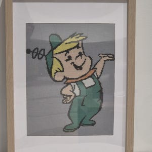 May include: Framed cross-stitch art featuring a cartoon character in green overalls, a yellow hat, and blonde hair. The character is set against a gray background and framed in a light wood frame. The artwork is a nostalgic piece.
